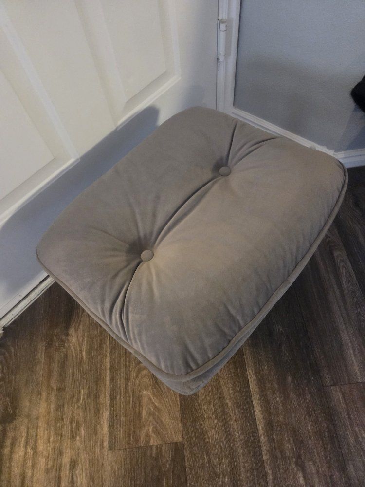 Tufted ottoman. Gray color. Has two buttons on top and piping around edge. Square shape.