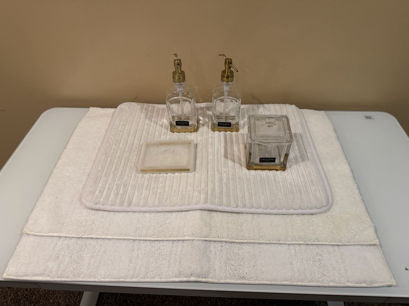Tahari Home 4-Piece Clear Glass & Gold Bathroom Vanity Set - Soap Dispenser, Tray, Jar and 3 white bathroom rugs.
